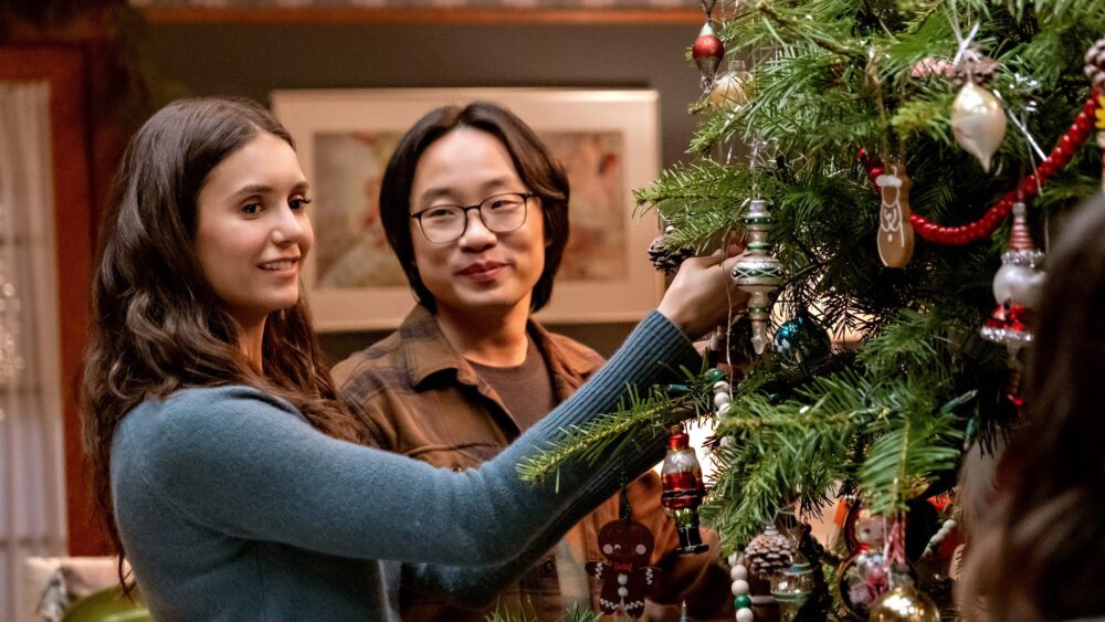 Two people(one Asian man, one white woman) stand smiling while trimming a Christmas tree